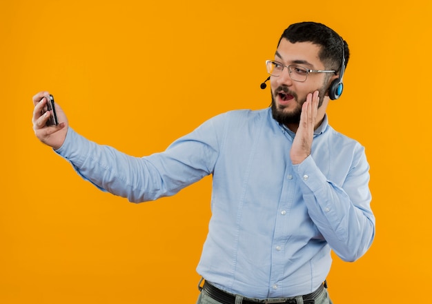 Jeune homme barbu à lunettes et chemise bleue avec des écouteurs avec microphone faisant selfie à l'aide de son smartphone souriant d'être surpris