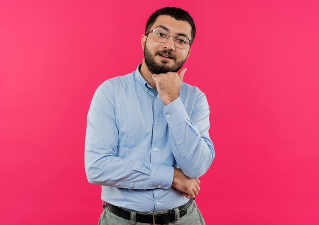 Jeune homme barbu à lunettes et chemise bleue avec la main sur le menton souriant et pensant