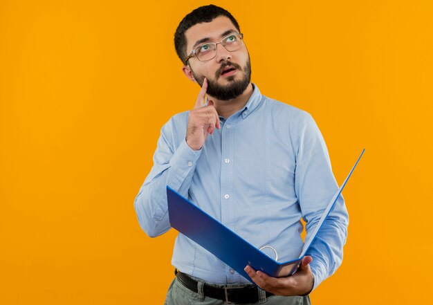 Jeune homme barbu à lunettes et chemise bleue tenant le dossier à la recherche avec une expression pensive pensée