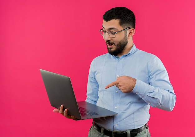 Jeune homme barbu à lunettes et chemise bleue tenant un ordinateur portable pointant avec le doigt sur le fait d'être surpris
