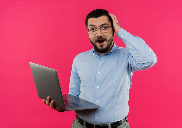 Jeune homme barbu à lunettes et chemise bleue tenant un ordinateur portable surpris et confus