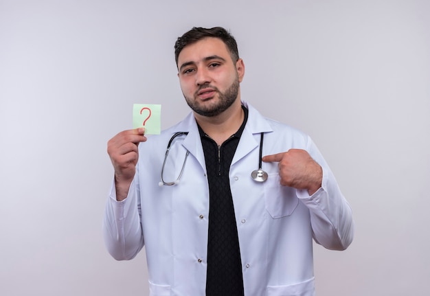 Jeune homme barbu médecin portant un manteau blanc avec stéthoscope tenant un papier de rappel avec point d'interrogation pointant avec l'index vers elle