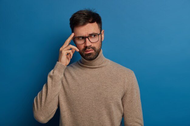 Un jeune homme barbu pensif garde le doigt sur la tempe, rappelle les informations nécessaires à l'esprit, porte des lunettes transparentes et un pull décontracté