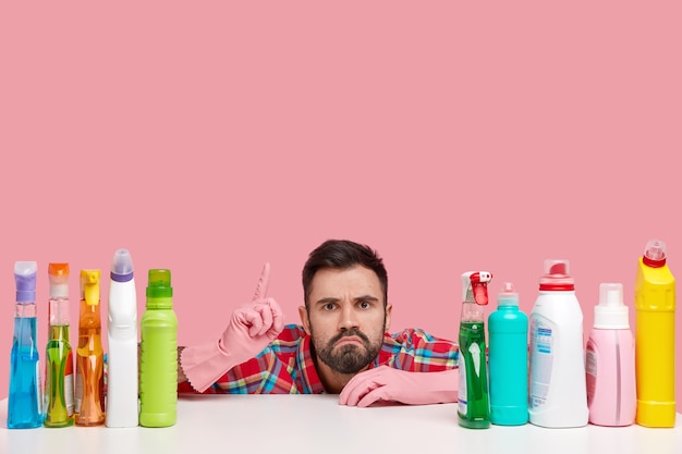 Un jeune homme barbu perplexe pointe vers le haut, a une expression mécontente, montre de la place pour nettoyer, utilise des détergents de haute qualité