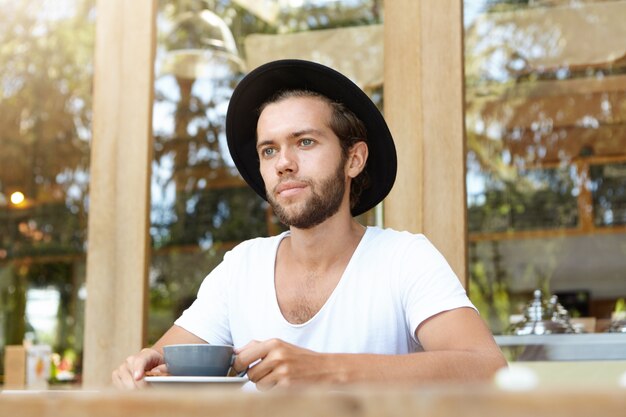 Jeune homme barbu de race blanche au chapeau à la mode ayant cappuccino, assis à table en bois avec mug au café en plein air