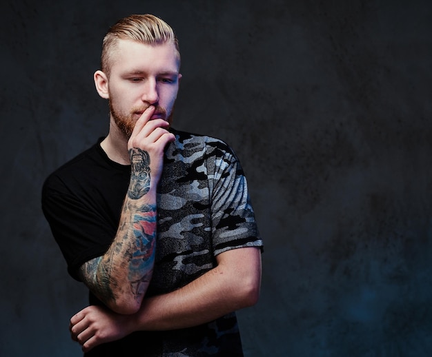 Photo gratuite jeune homme barbu rousse avec des tatouages sur les bras, vêtu d'un t-shirt noir sur fond gris foncé.