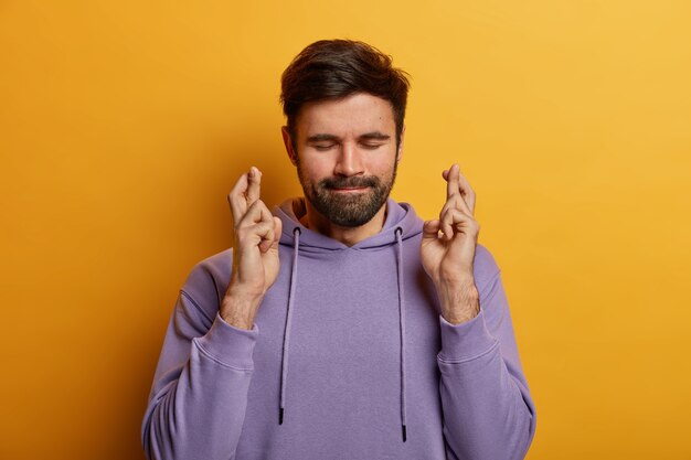 Un jeune homme barbu superstitieux croise les doigts pour avoir de la chance, espère que le souhait se réalise, ferme les yeux et presse les lèvres, a un grand désir ou rêve, porte un sweat-shirt violet, isolé sur un mur jaune