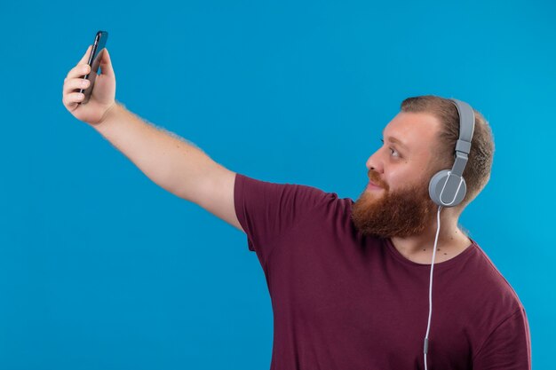 Jeune homme barbu en t-shirt marron avec un casque prenant selfie à l'aide de son smartphone