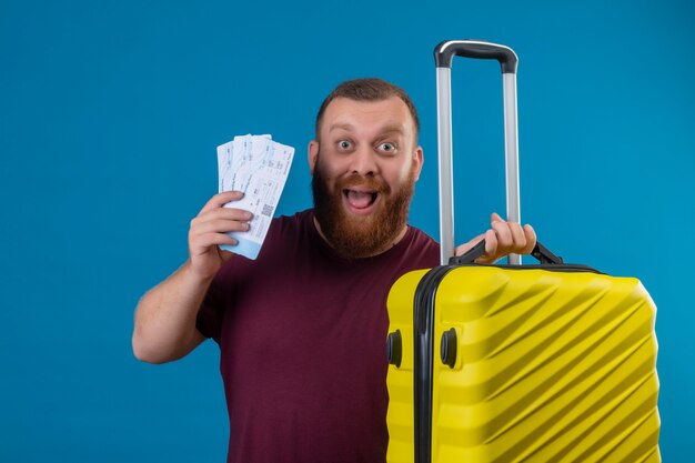 Jeune homme barbu en t-shirt marron tenant valise de voyage et billets d'avion à la sortie et heureux