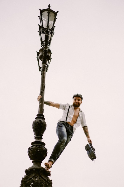 Jeune homme barbu tatoué. Un mec romantique en chemise blanche, casquette et bretelles se promène dans la ville. s'articule autour d'un lampadaire. Peaky Blinders. démodé, rétro.