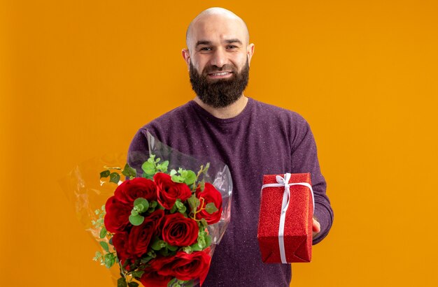 Jeune homme barbu tenant un bouquet de roses rouges et un présent souriant concept de jour de valentines confiant debout sur un mur orange