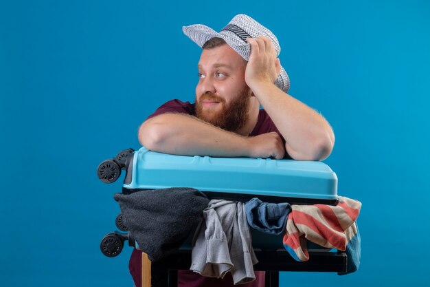 Jeune homme barbu voyageur en chapeau d'été avec valise pleine de vêtements optimiste et heureux souriant à côté avec un look de rêve