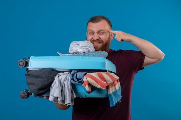 Jeune homme barbu voyageur tenant une valise pleine de vêtements pointant son temple en essayant de se souvenir