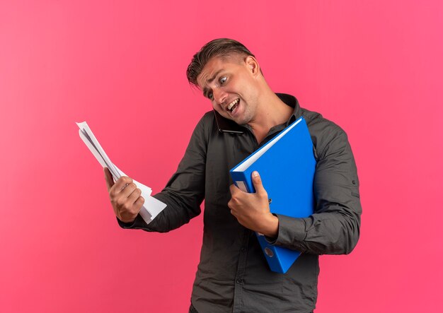 Jeune homme beau blond anxieux détient des feuilles de papier et des dossiers de fichiers parlant au téléphone isolé sur l'espace rose avec copie espace