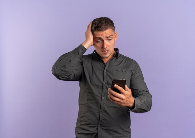 Jeune homme beau blond choqué tient la tête et regarde le téléphone isolé sur l'espace violet avec copie espace