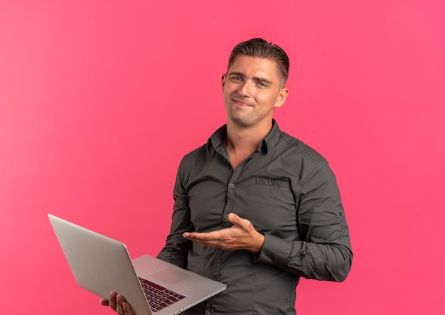 Jeune homme beau blond heureux tient et pointe sur un ordinateur portable isolé sur un espace rose avec espace de copie