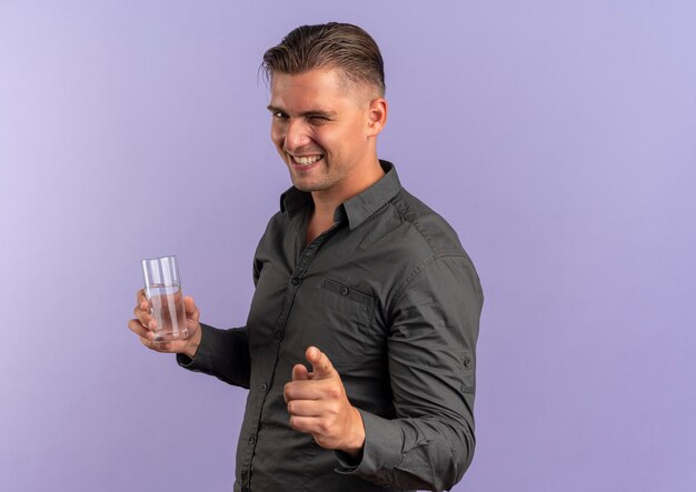 Jeune homme beau blond souriant clignote des yeux et pointe vers la caméra tenant un verre d'eau isolé sur fond violet avec espace de copie