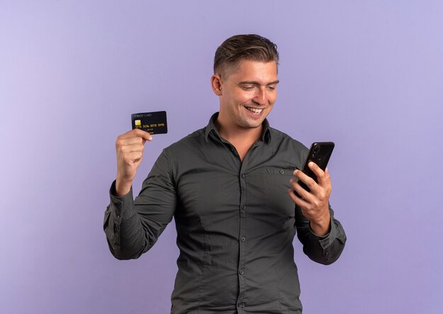 Jeune homme beau blond souriant détient la carte de crédit et regarde le téléphone