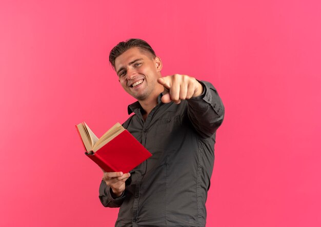 Jeune homme beau blond souriant tient le livre et les points à la caméra isolé sur fond rose avec copie espace