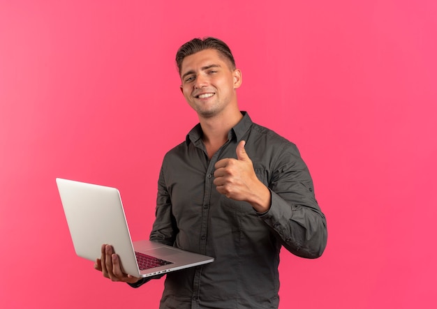 Jeune homme beau blond souriant tient un ordinateur portable et les pouces vers le haut isolé sur fond rose avec espace de copie