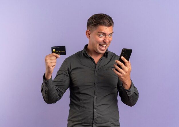 Jeune homme beau blonde surpris tient la carte de crédit et regarde le téléphone