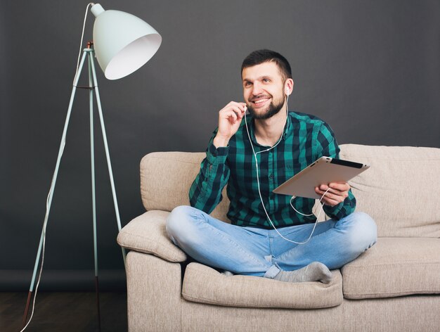 Jeune homme beau hipster assis sur un canapé à la maison tenant une tablette, écouter de la musique sur des écouteurs, parler en ligne, heureux, souriant, chemise à carreaux vert, loisirs, repos