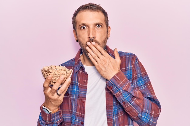 Photo gratuite jeune homme beau tenant des cornflakes couvrant la bouche avec la main choqué et peur de l'erreur expression surprise