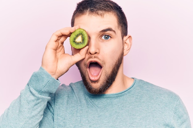 Photo gratuite jeune homme beau tenant une tranche de kiwi sur l'œil effrayé et étonné avec la bouche ouverte pour la surprise visage d'incrédulité