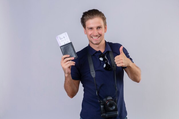 Jeune homme beau voyageur avec caméra tenant des billets d'avion regardant la caméra avec le sourire sur le visage heureux et positif montrant les pouces vers le haut debout sur fond blanc