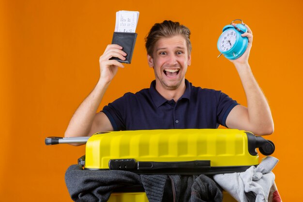 Jeune homme beau voyageur debout avec une valise pleine de vêtements tenant un réveil et des billets d'avion surpris et sorti avec une expression de confusion sur fond orange