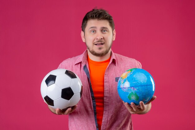 Jeune homme beau voyageur tenant un ballon de football et un globe avec une expression de confusion