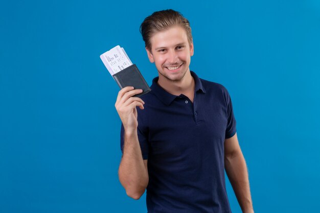 Jeune homme beau voyageur tenant des billets d'avion heureux et positif regardant la caméra avec un grand sourire sur le visage debout sur fond bleu