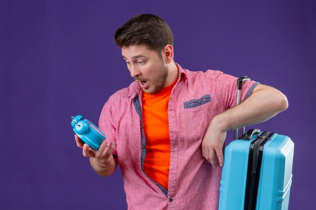 Jeune homme beau voyageur tenant une valise bleue regardant réveil dans sa main