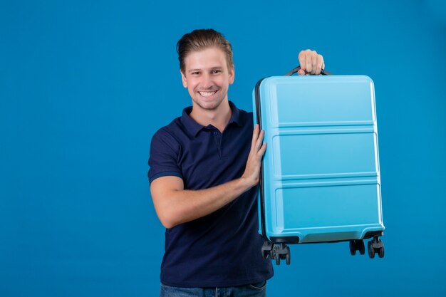 Jeune homme beau voyageur tenant valise regardant la caméra en souriant joyeusement avec un visage heureux debout sur fond bleu