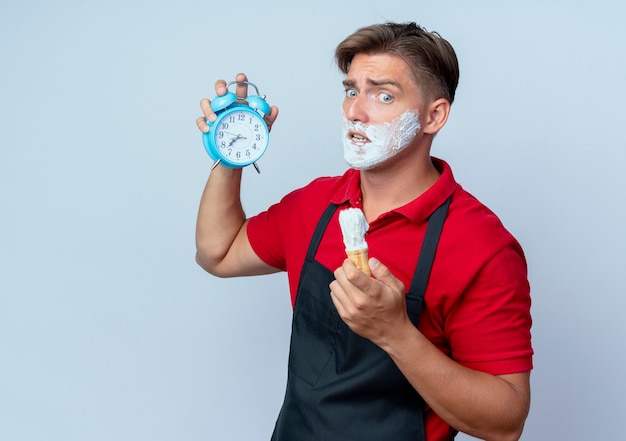 Jeune homme blond anxieux coiffeur en uniforme visage enduit de mousse à raser tenant un réveil et un blaireau à la bas isolé sur un espace blanc avec espace copie