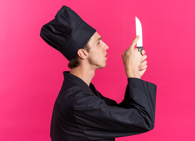 Photo gratuite jeune homme blond concentré cuisinier en uniforme de chef et casquette debout en vue de profil tenant regardant et touchant le couteau avec le doigt