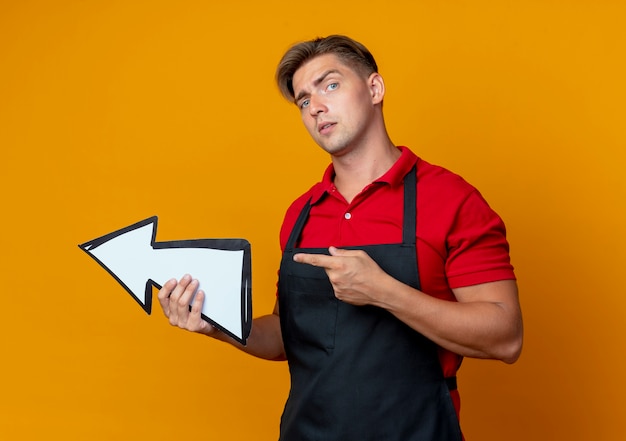 Jeune homme blond confiant barbier en uniformholds direction mark et points sur le côté isolé sur l'espace orange avec copie espace