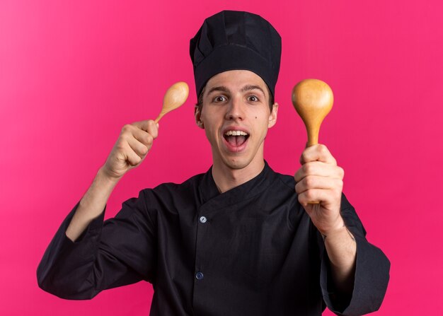 Jeune homme blond excité cuisinier en uniforme de chef et casquette montrant des cuillères regardant la caméra isolée sur un mur rose