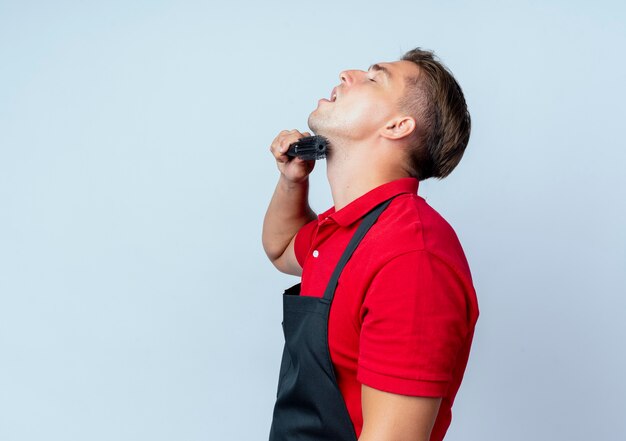 Photo gratuite jeune homme blond heureux coiffeur en uniforme se tient sur le côté peignant le menton isolé sur un espace blanc avec copie