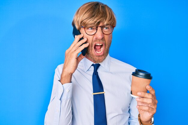 Jeune homme blond parlant sur le smartphone et buvant une tasse de café en colère et fou criant frustré et furieux, criant de colère. rage et concept agressif.
