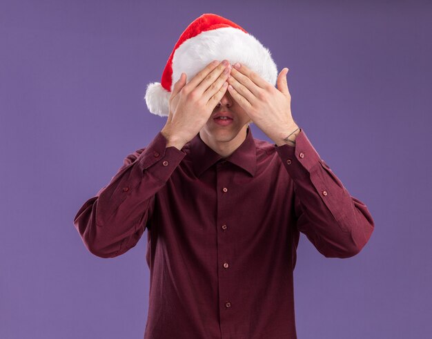 Jeune homme blond portant bonnet de Noel et lunettes couvrant ses yeux avec les mains isolées sur fond violet