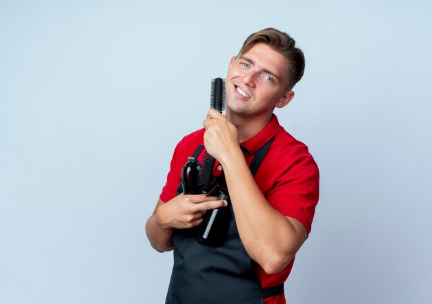 Jeune homme blond souriant coiffeur en uniforme détient des outils de coiffeur isolés sur un espace blanc avec copie espace