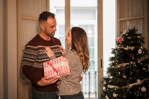 Jeune homme avec une boîte cadeau embrassant une dame joyeuse près de sapin de Noël