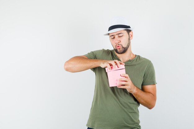 Jeune homme boîte-cadeau d'ouverture soigneusement en t-shirt vert et chapeau et à la curiosité