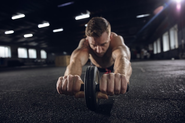 Jeune homme en bonne santé, athlète faisant des exercices avec le rouleau dans la salle de gym. Modèle masculin célibataire pratiquant durement et entraînant le haut de son corps. Concept de mode de vie sain, sport, fitness, musculation, bien-être.