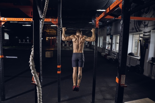 Jeune homme en bonne santé, athlète faisant des exercices, tractions en salle de sport. Modèle caucasien unique pratiquant dur, entraînant le haut de son corps. Concept de mode de vie sain, sport, fitness, musculation, bien-être.