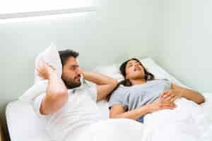 Photo gratuite jeune homme bouleversé couvrant ses oreilles avec un oreiller et se sentant en colère contre sa petite amie ronflante en position couchée dans son lit