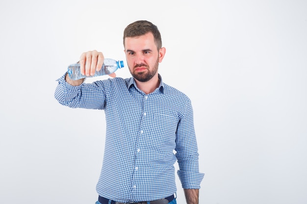 Jeune homme bouteille d'eau en plastique inclinable en chemise, jeans et à la vue de face, hésitant.