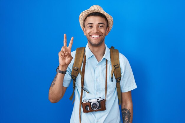 Jeune homme brésilien tenant un appareil photo vintage montrant et pointant vers le haut avec les doigts numéro deux tout en souriant confiant et heureux