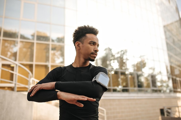 Jeune homme brun bouclé fait des exercices pour les bras à l'extérieur Beau mec à la peau foncée en tenue noire travaillant dans la rue